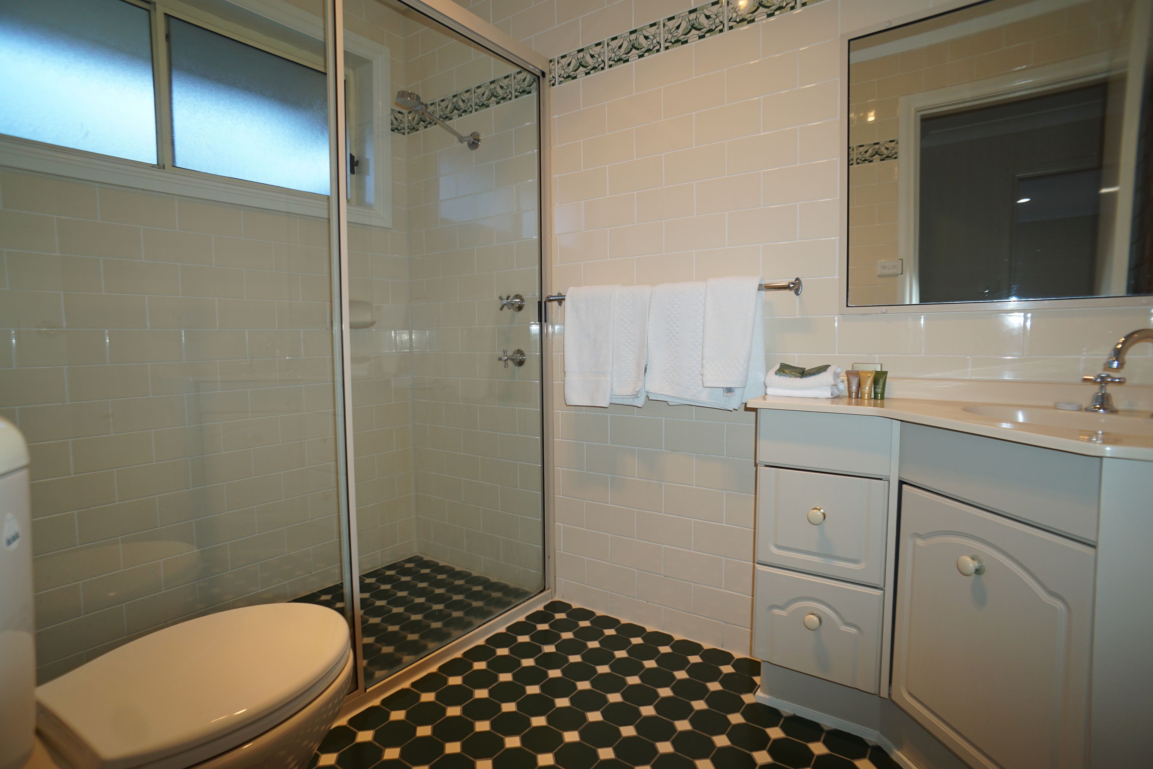 Un baño con cabina de ducha de vidrio, paredes de azulejos blancos, suelo a cuadros blanco y negro y un lavabo blanco con un espejo encima.