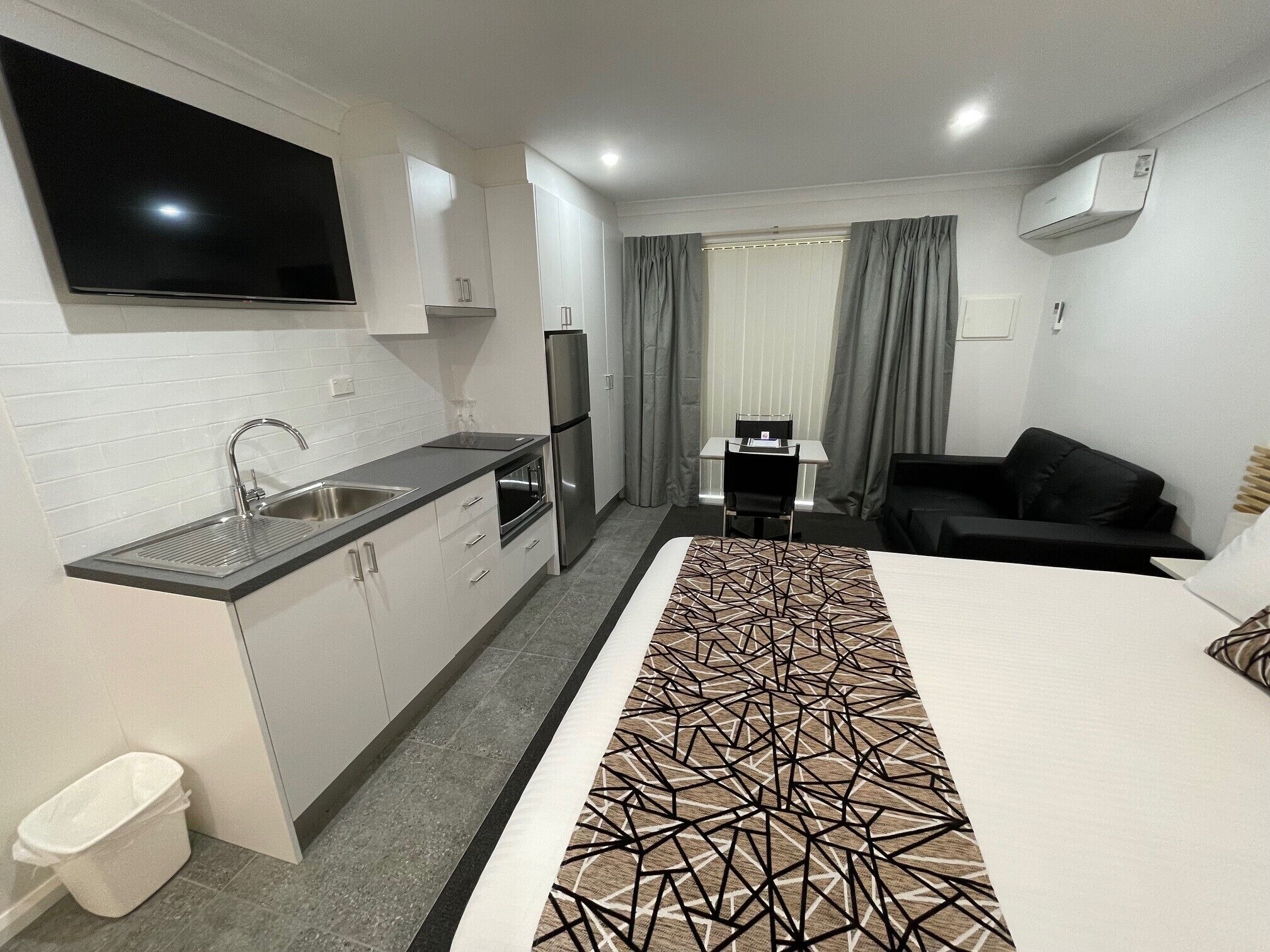 Ein modernes Hotelzimmer mit Küchenzeile, einem Bett mit gemustertem Überwurf, einer Couch und einem an der Wand montierten Fernseher.
