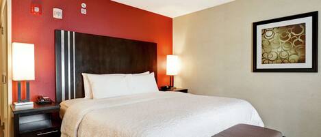 Une chambre d’hôtel avec un grand lit, une table de chevet avec une lampe, un bureau avec un téléphone et une œuvre d’art encadrée au mur.