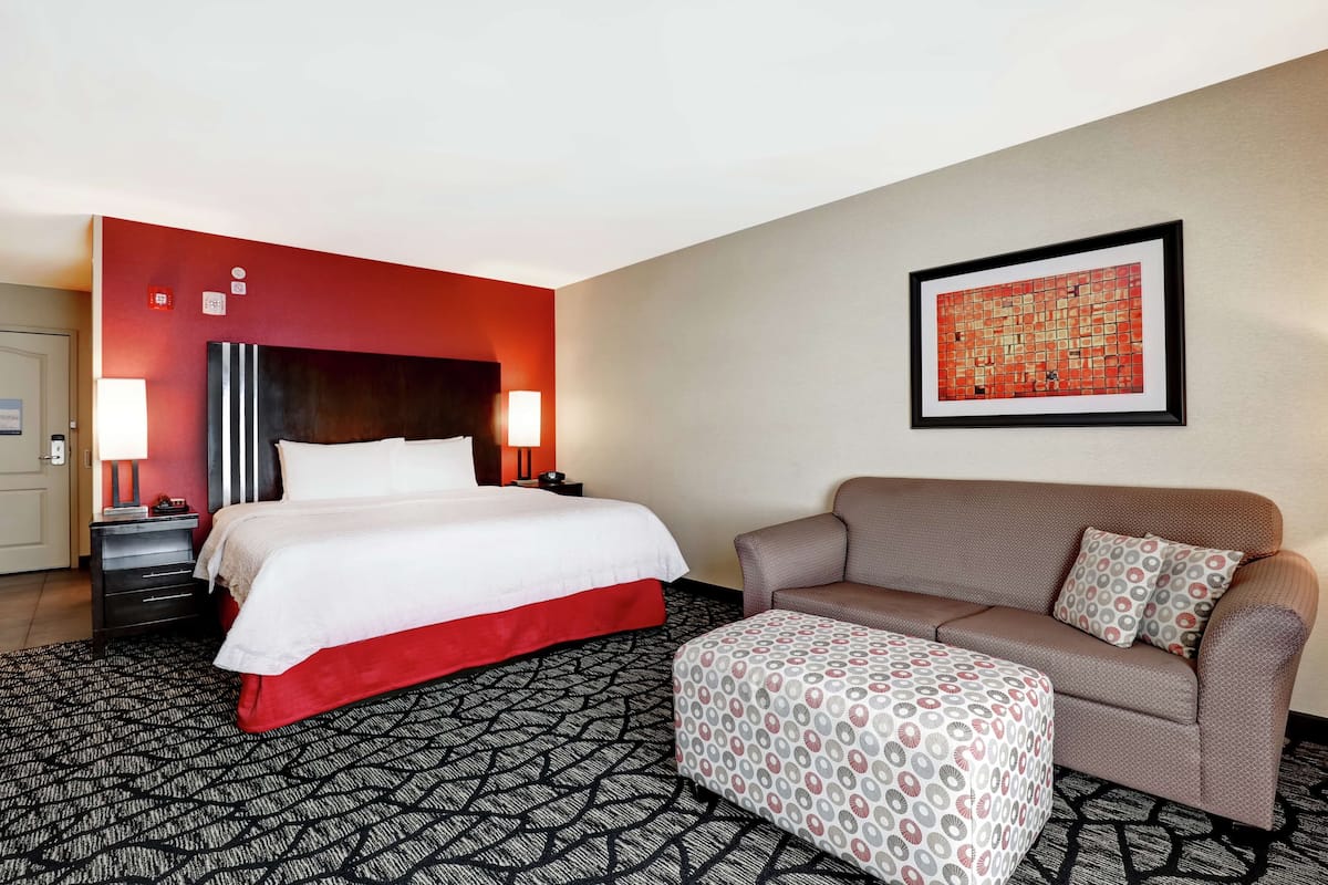 Quarto de hotel com cama, sofá, um banquinho estampado, mesa de cabeceira, luminária e uma obra de arte emoldurada na parede.