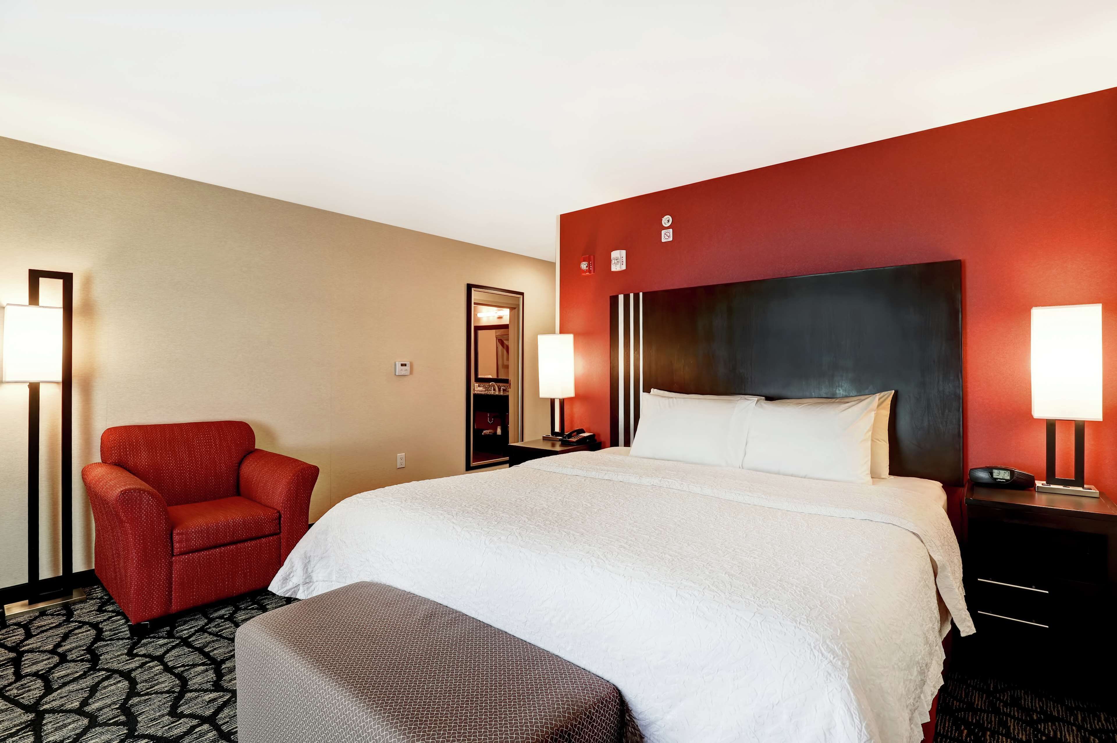 Habitación de hotel con una cama grande, un sillón rojo, una mesita de noche con lámpara y un espejo en la pared.