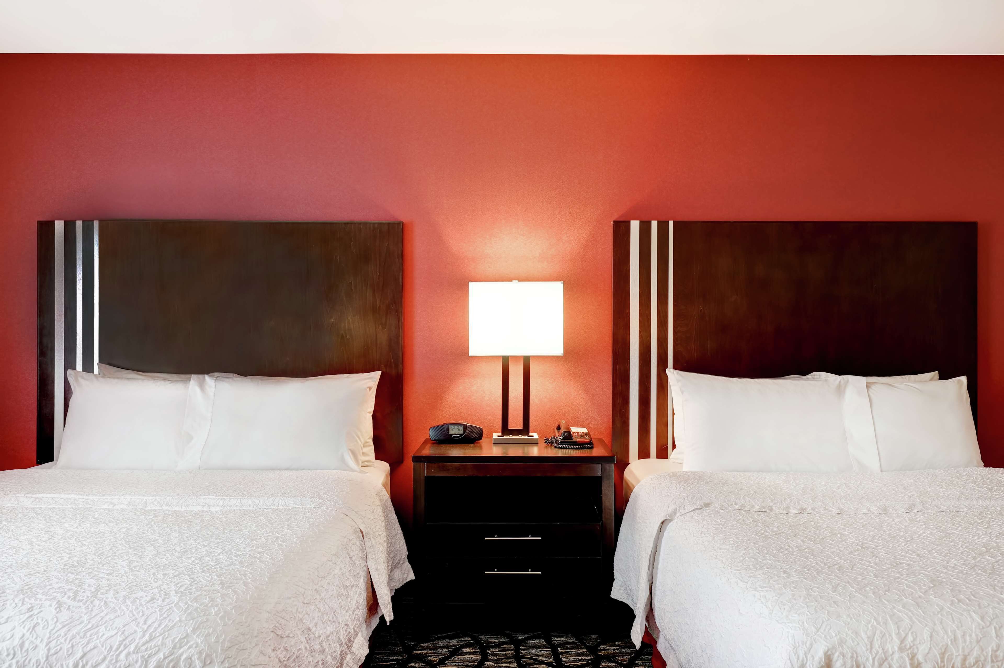 A hotel room with two beds, a nightstand with a lamp, and a red wall.