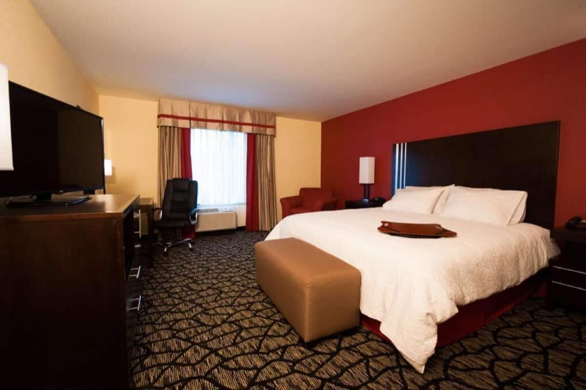 Quarto de hotel com uma cama grande, uma escrivaninha com televisão, uma cadeira e um pequeno banquinho.