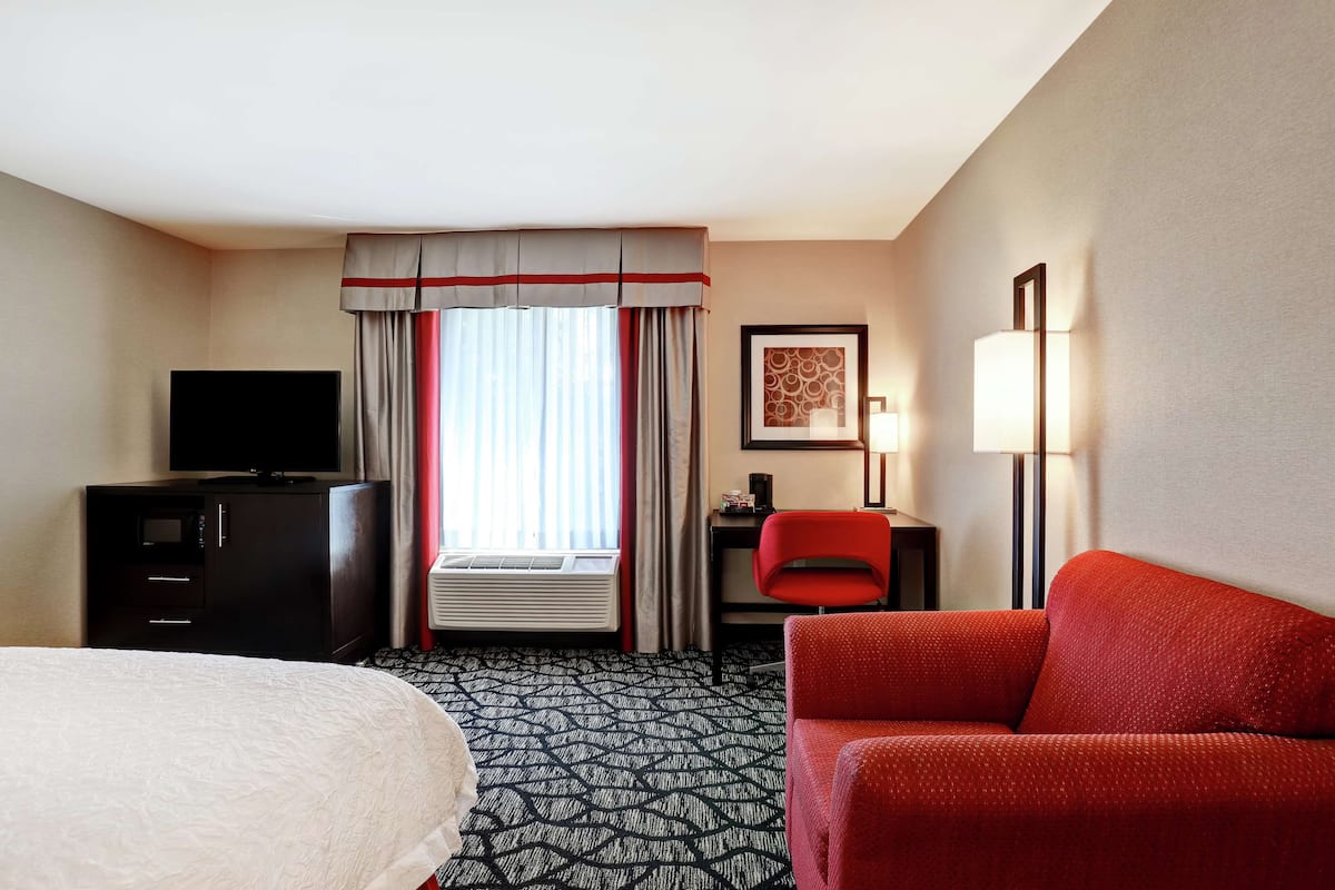 Quarto de hotel com cama, escrivaninha com cadeira, televisão, abajur e janela com cortinas.