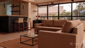 A modern living room with a sofa, two bar stools, a glass coffee table, and a kitchen area with a bar counter. - Rede Andrade SLZ (São Luís)