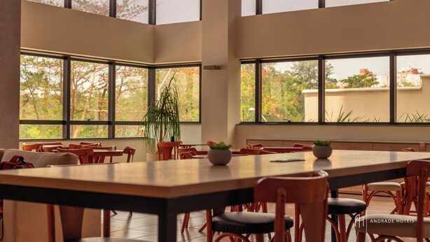 A spacious dining area with a long table and multiple chairs, large windows offering views of trees and buildings, and potted plants on the table. - Rede Andrade SLZ (São Luís)