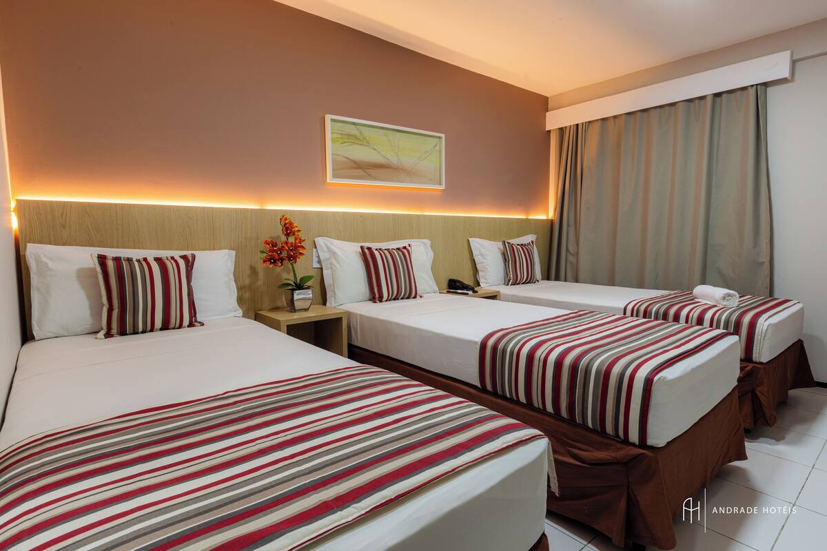 A hotel room with three beds, each with striped bedding, a nightstand with a vase of flowers, a painting on the wall, and a window with curtains.