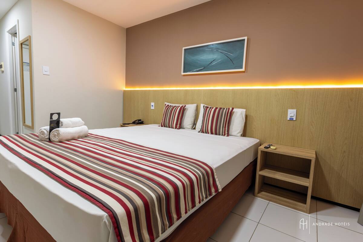 A hotel room with a large bed, striped bedding, a bedside table, a nightstand, a painting on the wall, and a door.