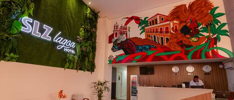 A hotel reception area with a neon sign reading "SLZ Lagoa Hotel", a mural of a horse and buildings, and a reception desk with a clock and a computer.