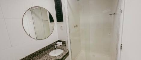 A bathroom with a granite countertop, a round mirror, a glass shower enclosure, and white tiled walls.