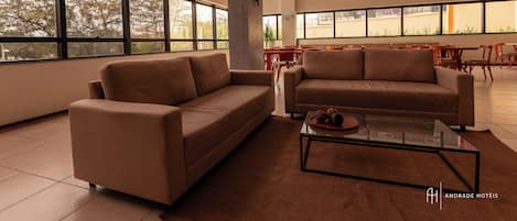 A spacious hotel lobby with large windows, two sofas, a glass coffee table, and a red dining area in the background.