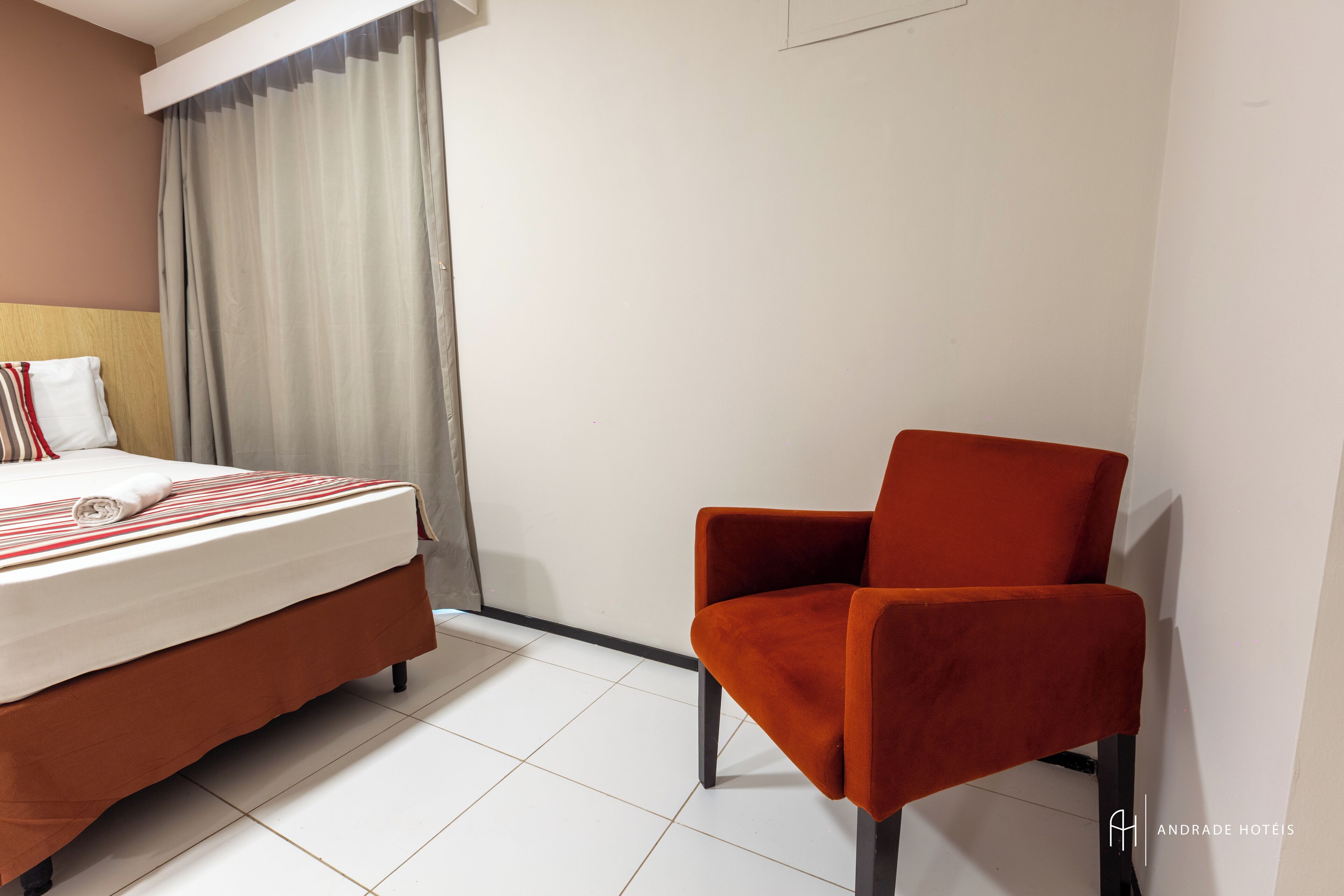 A hotel room with a bed, a red chair, and a white curtain.