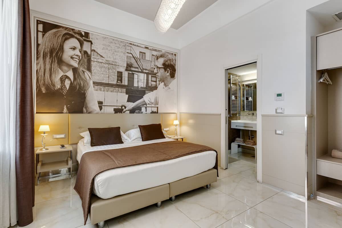 A hotel room with a large bed, two bedside tables with lamps, a bathroom with a mirror and shelves, and a wall with a large black-and-white photograph.
