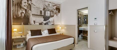 A hotel room with a large bed, two bedside tables with lamps, a bathroom with a mirror and shelves, and a wall with a large black-and-white photograph.