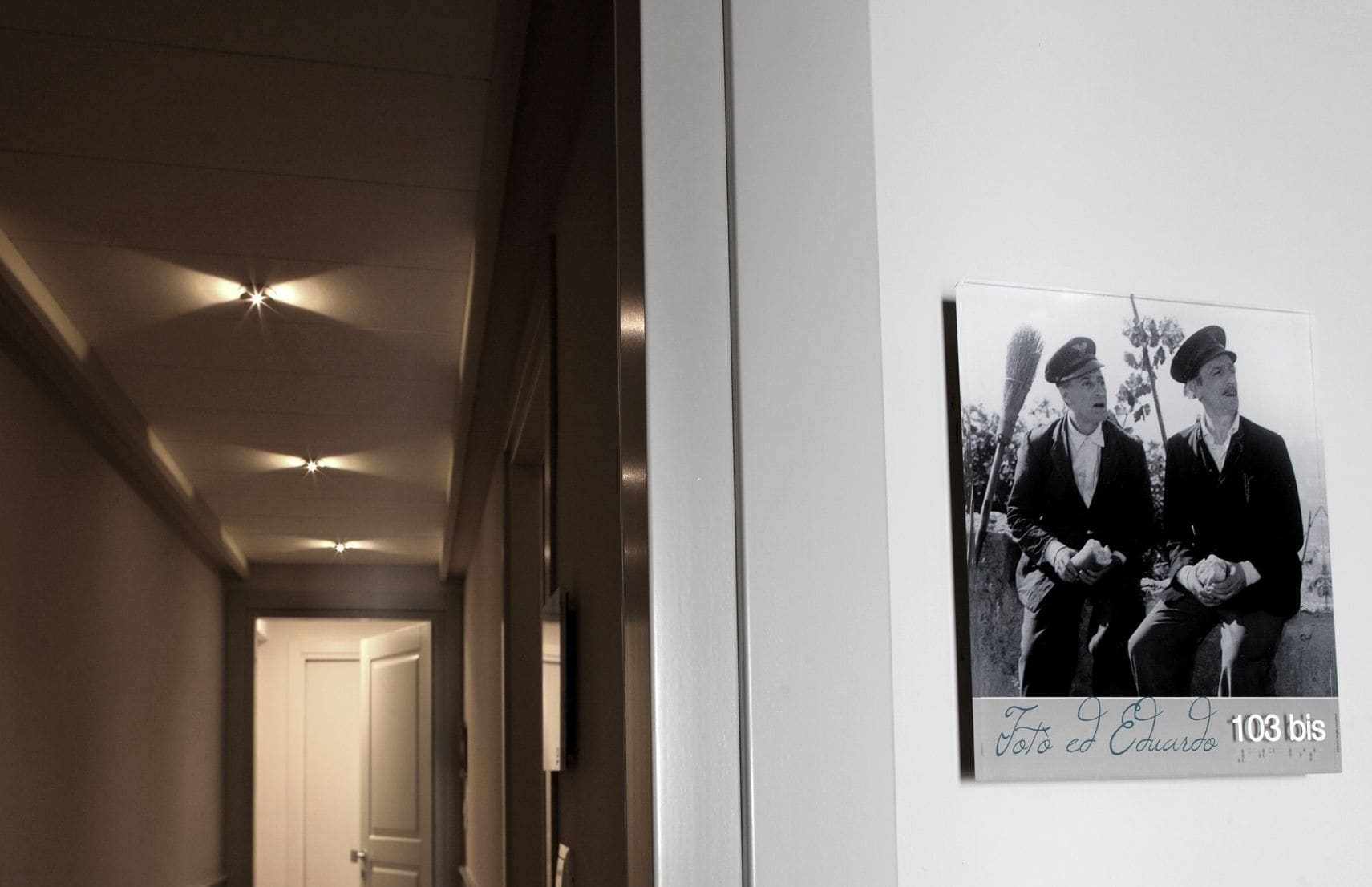 Un pasillo con una fotografía blanco y negro enmarcada en la pared, una puerta al final e iluminación empotrada en el techo.