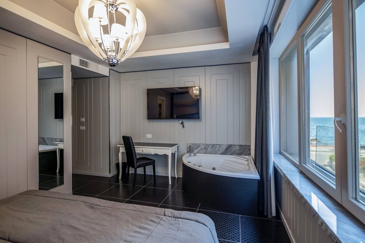 A modern bedroom with a large bed, a desk, a chair, a TV, and a bathtub.