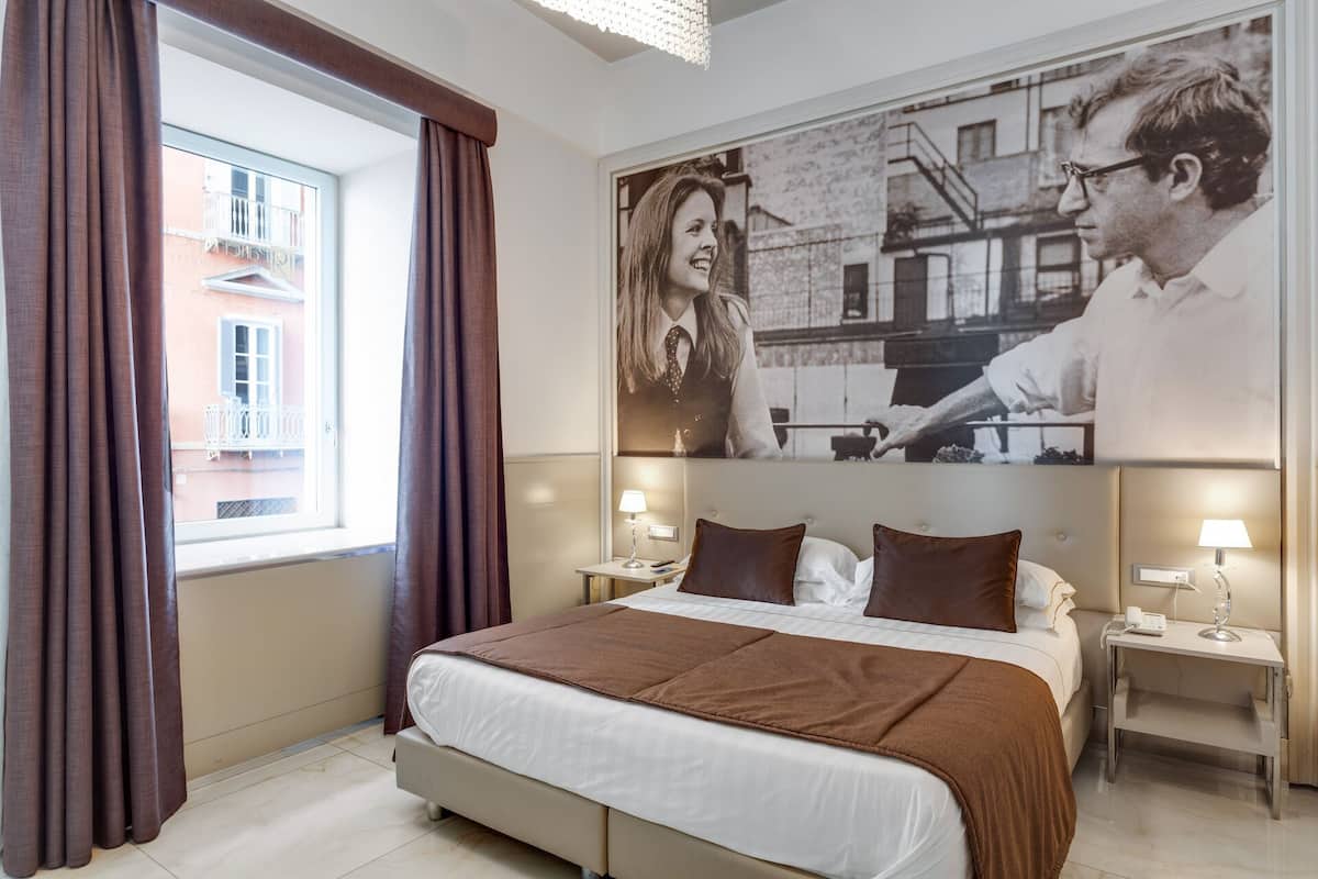 A hotel room with a large bed, two bedside tables, a window with curtains, and a wall with a large black and white photograph.