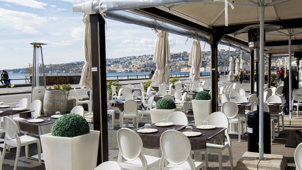 An outdoor cafe with white chairs and tables, a view of the city, and a covered area with umbrellas. - Partenope Relais (Naples)