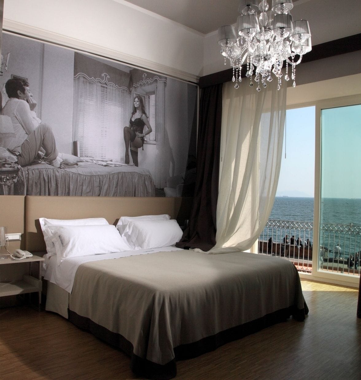 Camera d'albergo con un letto grande, una scrivania con il telefono, un lampadario e vista sul mare da un balcone.