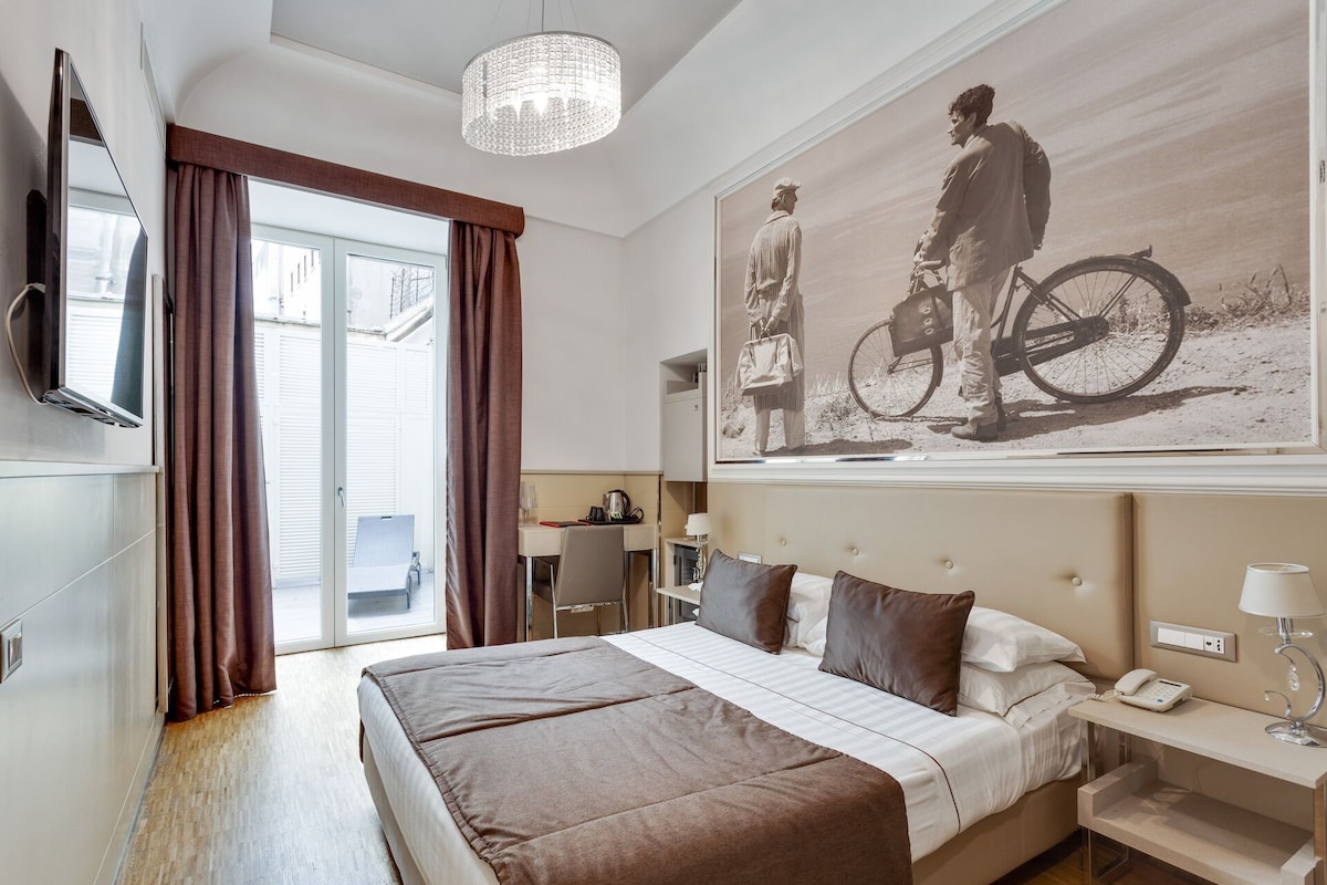 A hotel room with a bed, a desk, a chair, a TV, a chandelier, and a large painting.