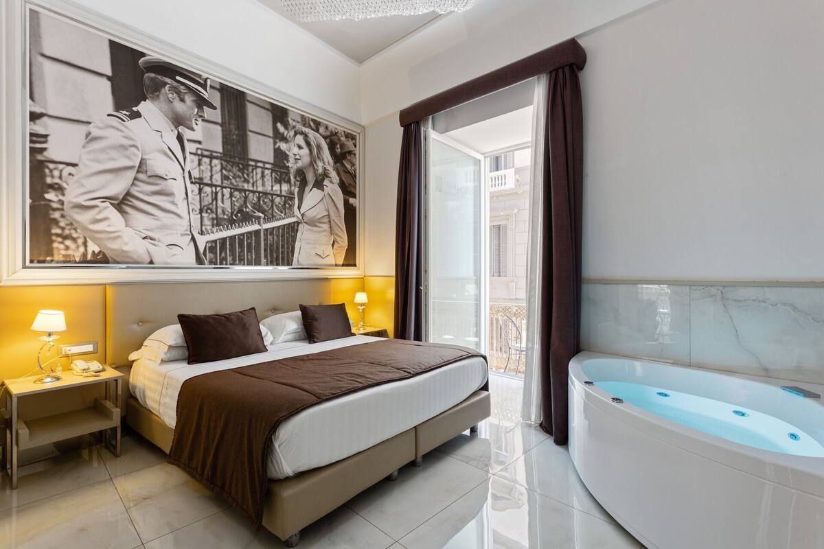 A hotel room with a large bed, a bedside table with a lamp, a large framed photograph on the wall, and a bathtub.