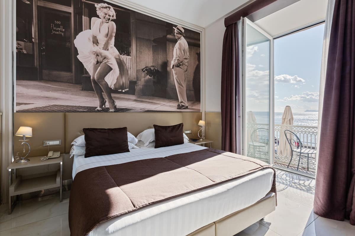 A hotel room with a large bed, a nightstand, a lamp, and a view of the sea through a balcony.