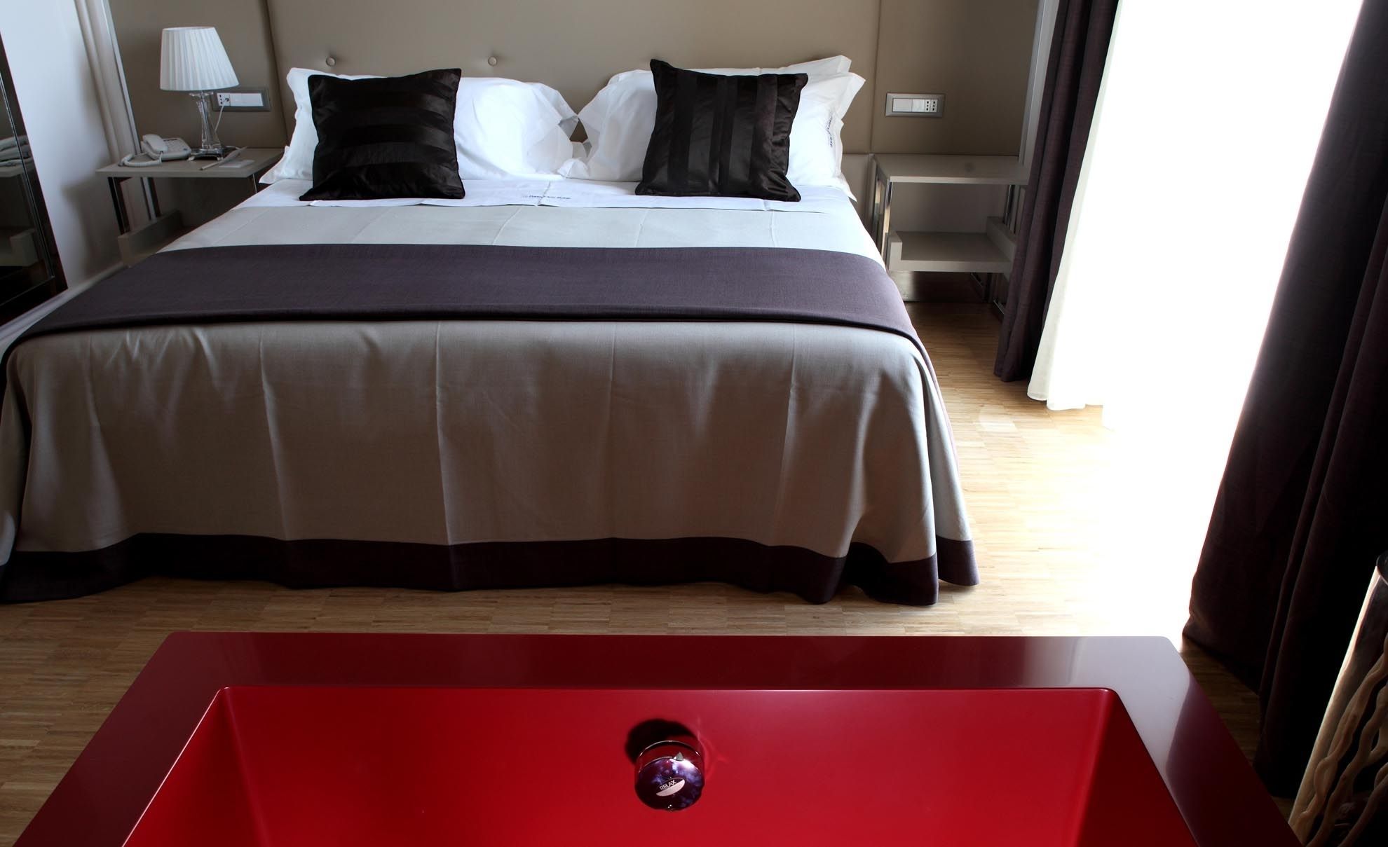 A hotel room with a bed, a red table, and a nightstand with a lamp.