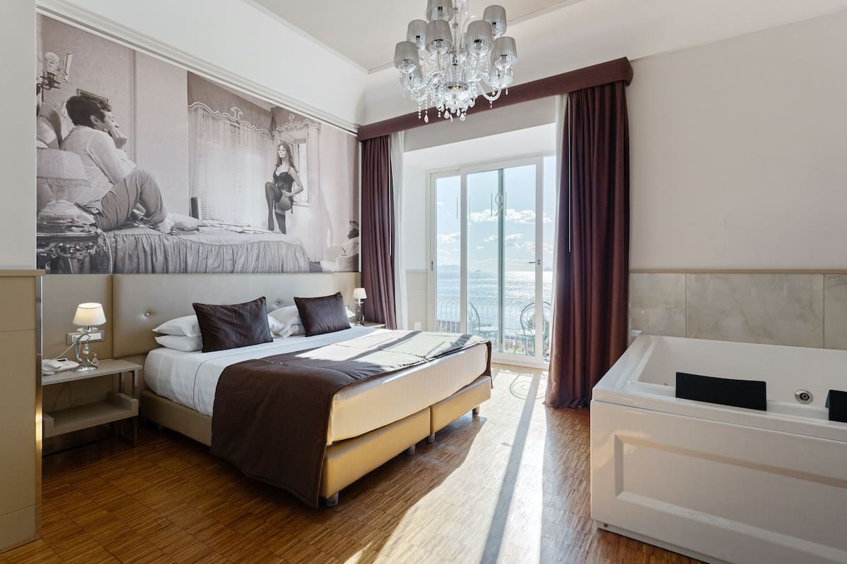 A hotel room with a large bed, a separate sitting area, a bathtub, and a balcony with a view.