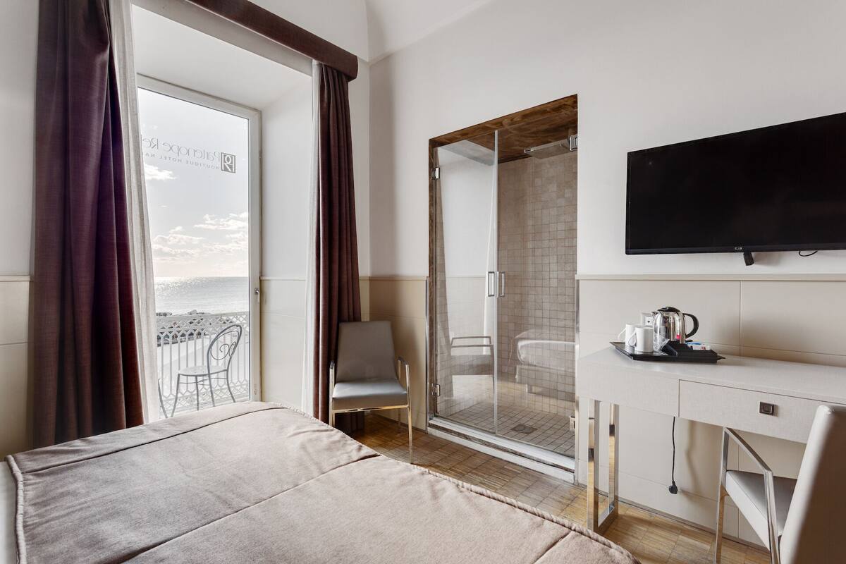A hotel room with a bed, a desk with a TV, a shower area, and a view of the sea.
