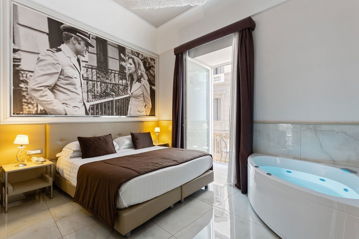 A hotel room with a bed, a large window, a jacuzzi, and a framed picture on the wall.