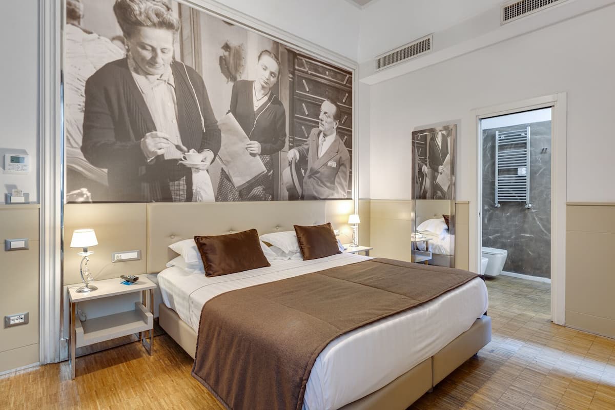 A modern hotel room with a large bed, bedside tables with lamps, a large wall-mounted picture, and a view of the outside through a door.