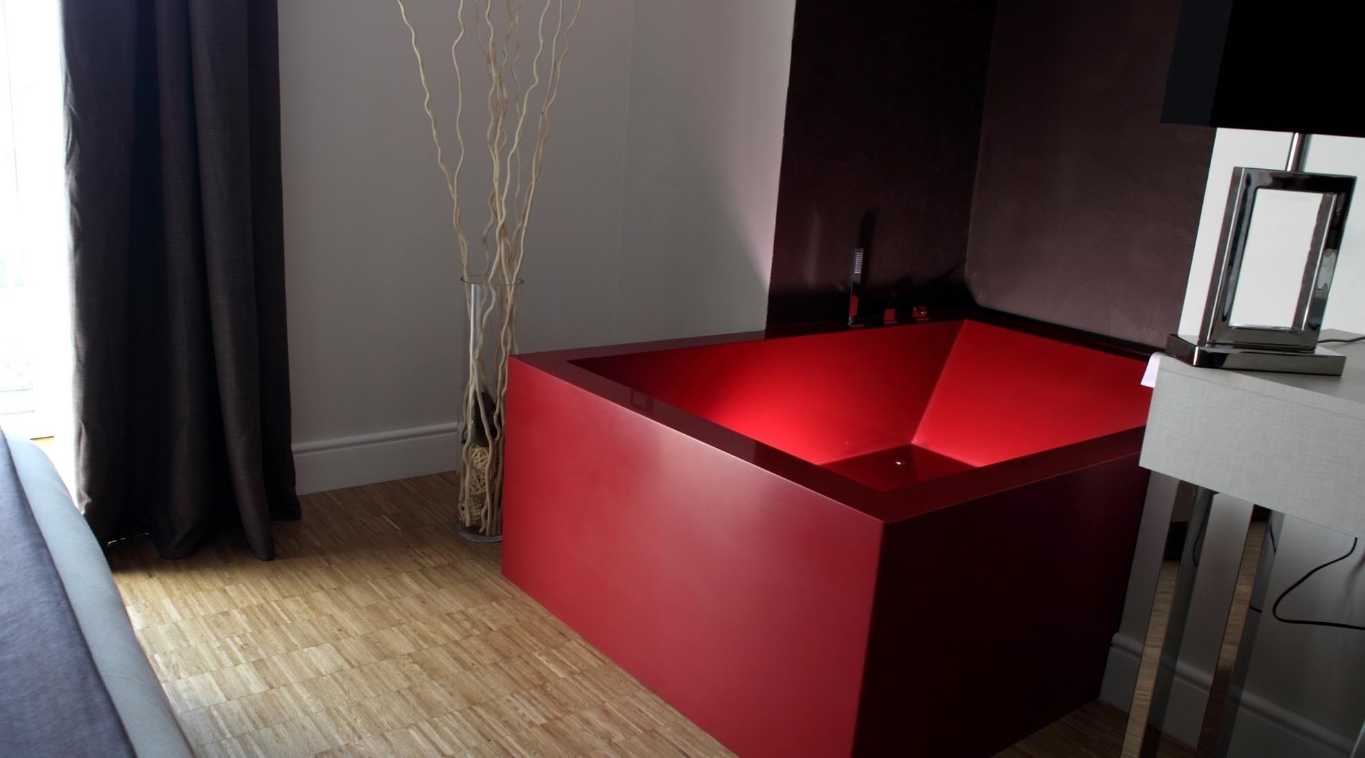 Un baño moderno con una bañera roja, un florero de vidrio con ramas y un espejo en la pared.