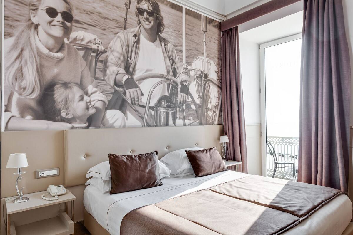 A hotel room with a large bed, a headboard with a mounted artwork, a bedside table with a lamp, and a view of the outdoors through a window with curtains.