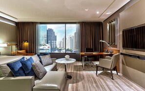 A hotel room with a sofa, a small round table, a desk with a chair, a TV, and a view of the cityscape. - Hilton Sukhumvit Bangkok (Bangkok)