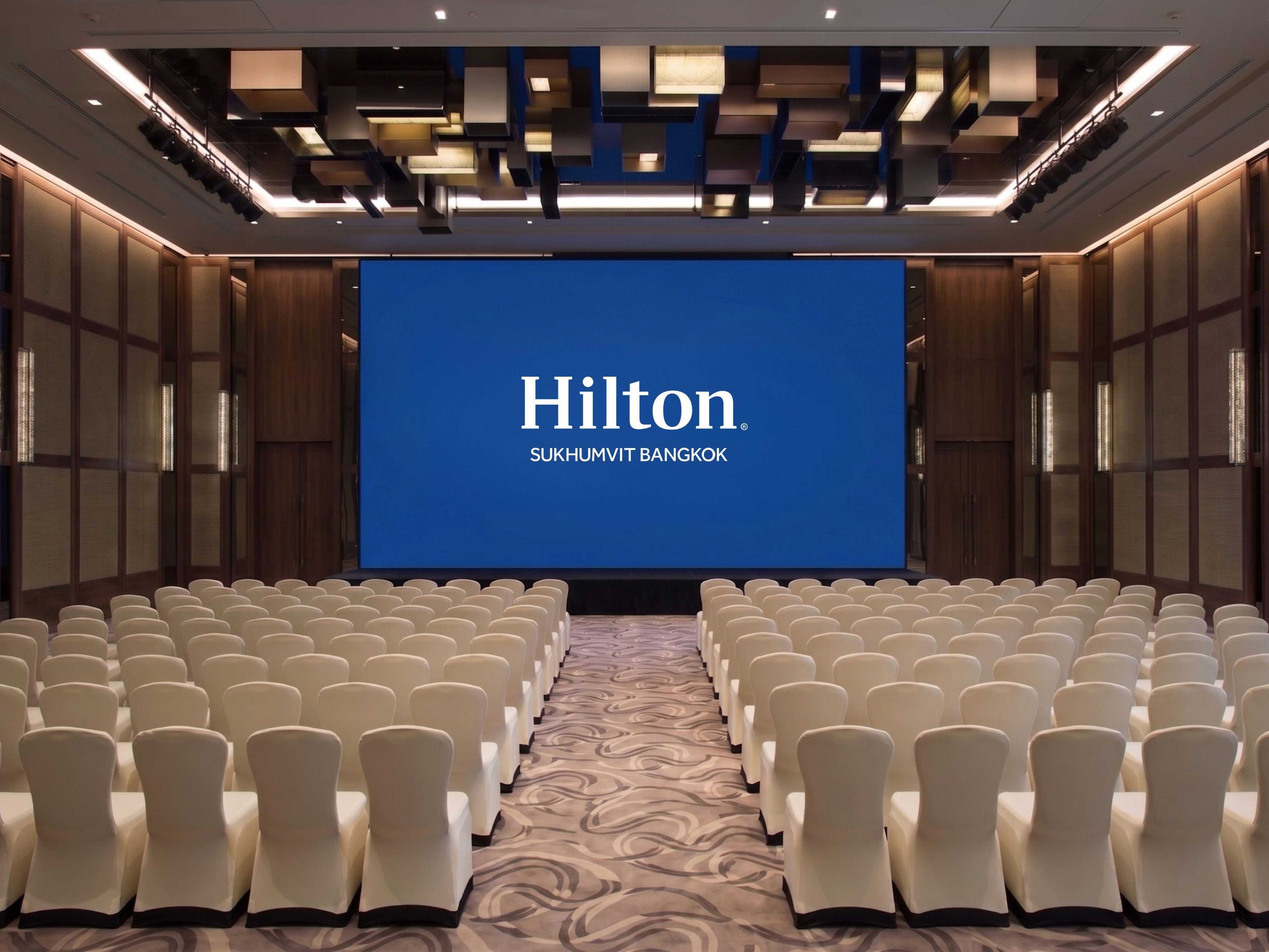 A conference room with a large screen displaying 'Hilton Sukhumvit Bangkok' and rows of white chairs.