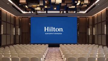 A conference room with a large screen displaying 'Hilton Sukhumvit Bangkok' and rows of white chairs.