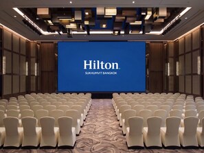 Uma sala de conferências com uma tela grande exibindo 'Hilton Sukhumvit Bangkok' e fileiras de cadeiras brancas.