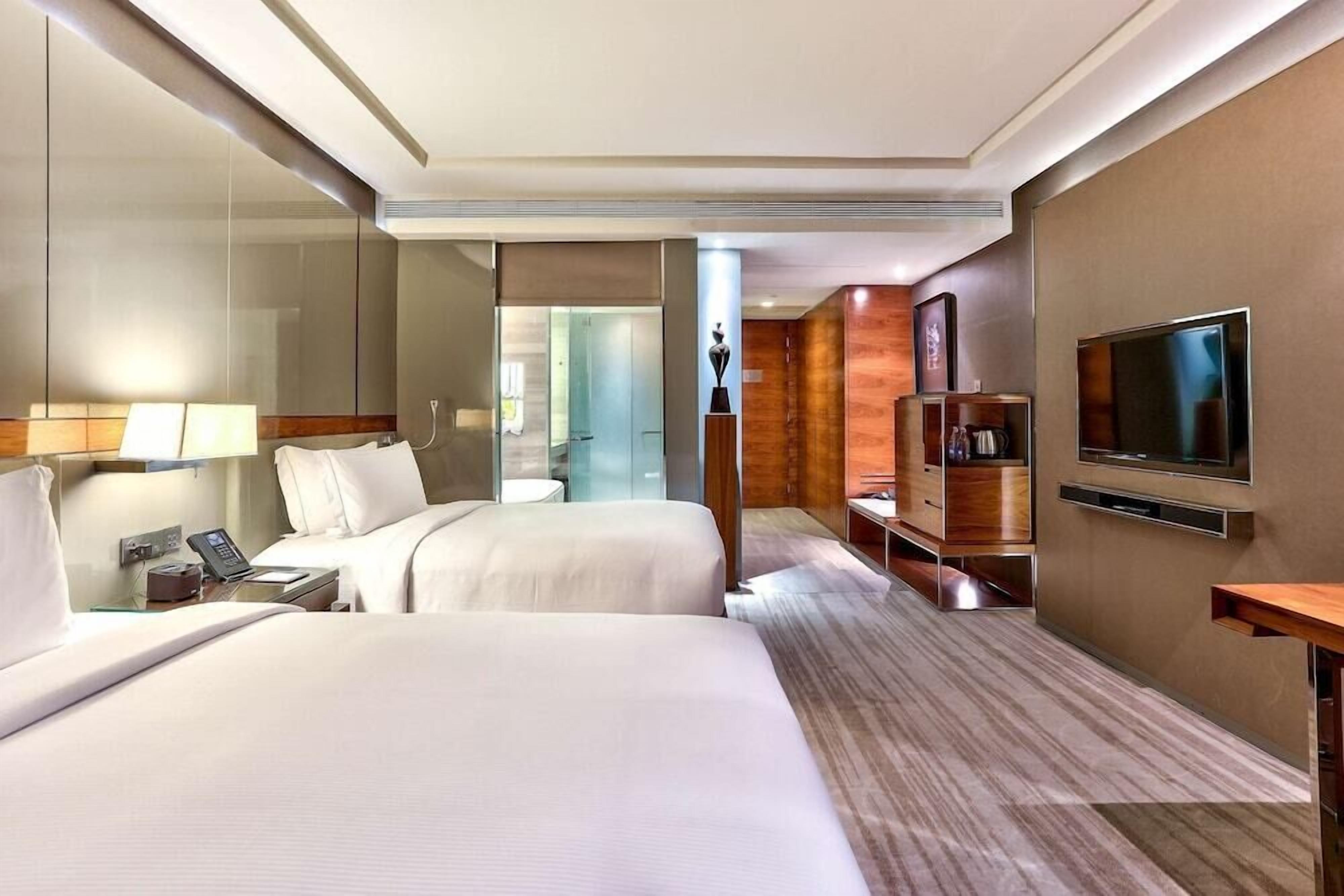 Une chambre d’hôtel avec deux lits, une télévision, un bureau avec un téléphone et une salle de bain visible à travers une porte en verre.