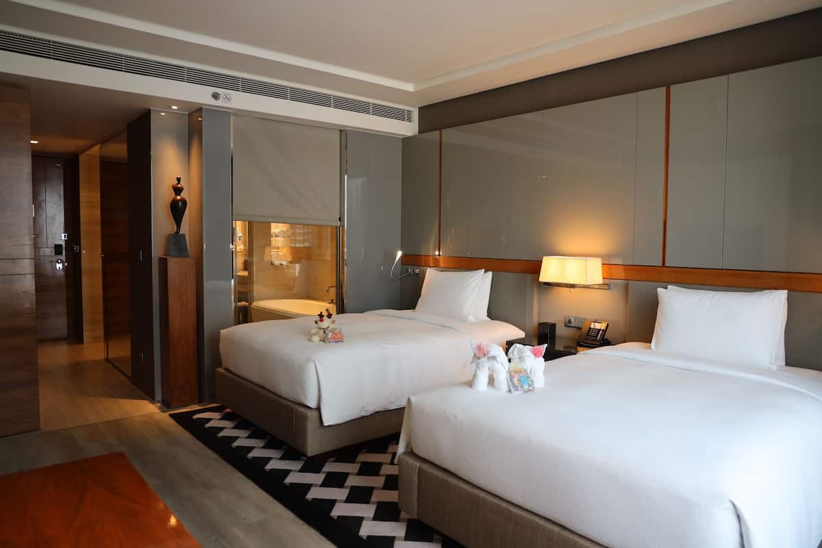Une chambre d’hôtel avec deux lits, un grand miroir, une table de chevet et une salle de bain visible à travers une porte ouverte.