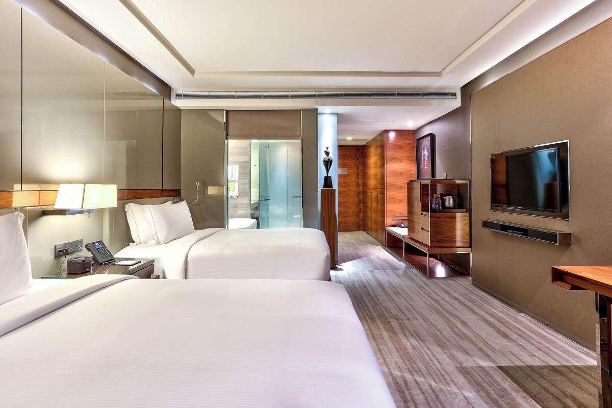 Une chambre d’hôtel avec deux lits, une télévision, un bureau avec un téléphone et une salle de bain visible à travers une porte en verre.