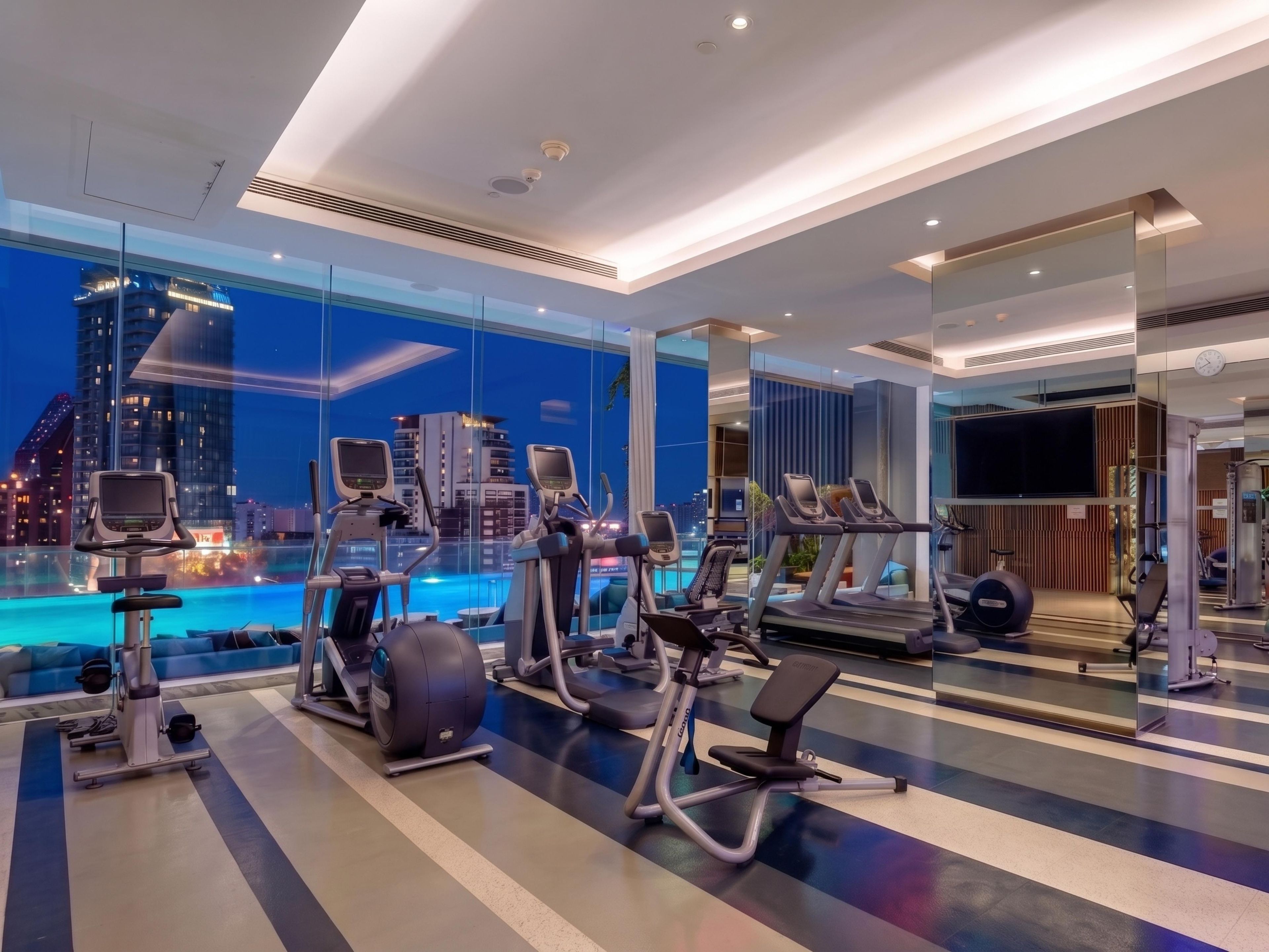 A modern gym with various exercise equipment, a large window overlooking a cityscape, and a pool area visible through the glass.