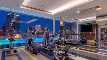 A modern gym with various exercise equipment, a large window overlooking a cityscape, and a pool area visible through the glass.