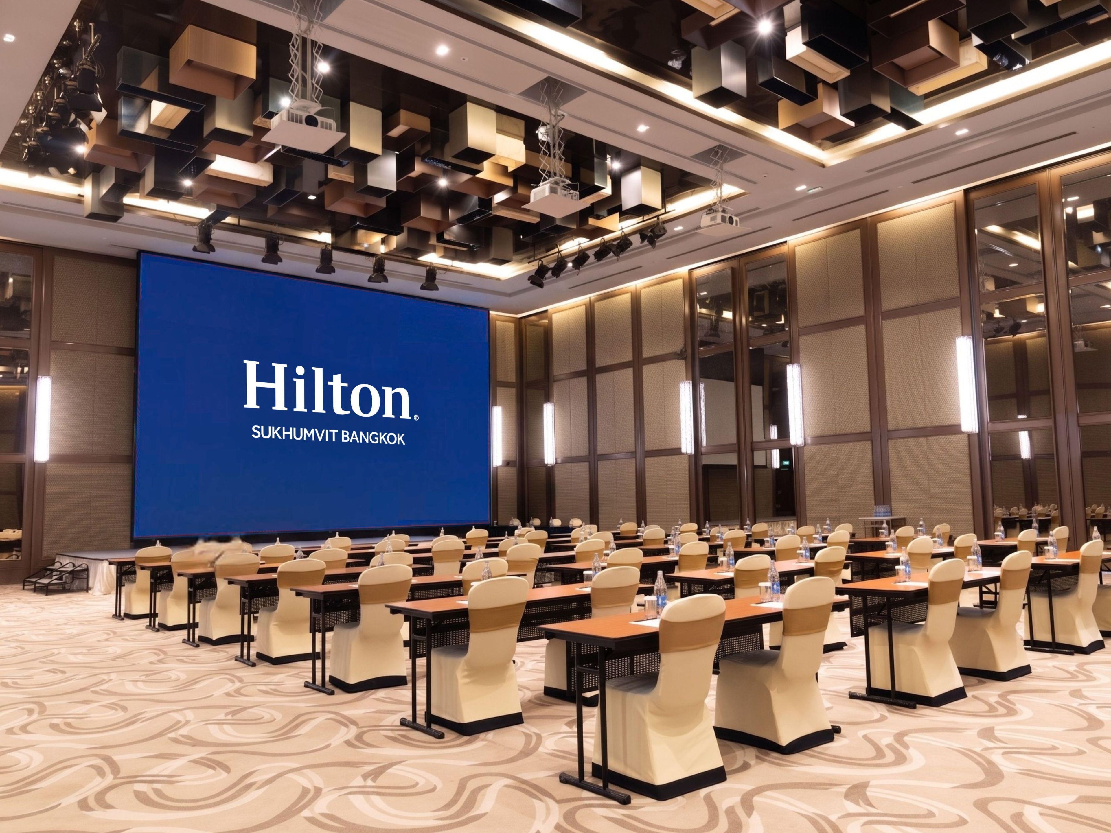 A conference room with a large screen displaying 'Hilton Sukhumvit Bangkok' and rows of tables with chairs.