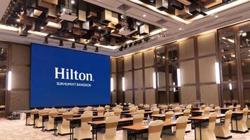 A conference room with a large screen displaying 'Hilton Sukhumvit Bangkok' and rows of tables with chairs.
