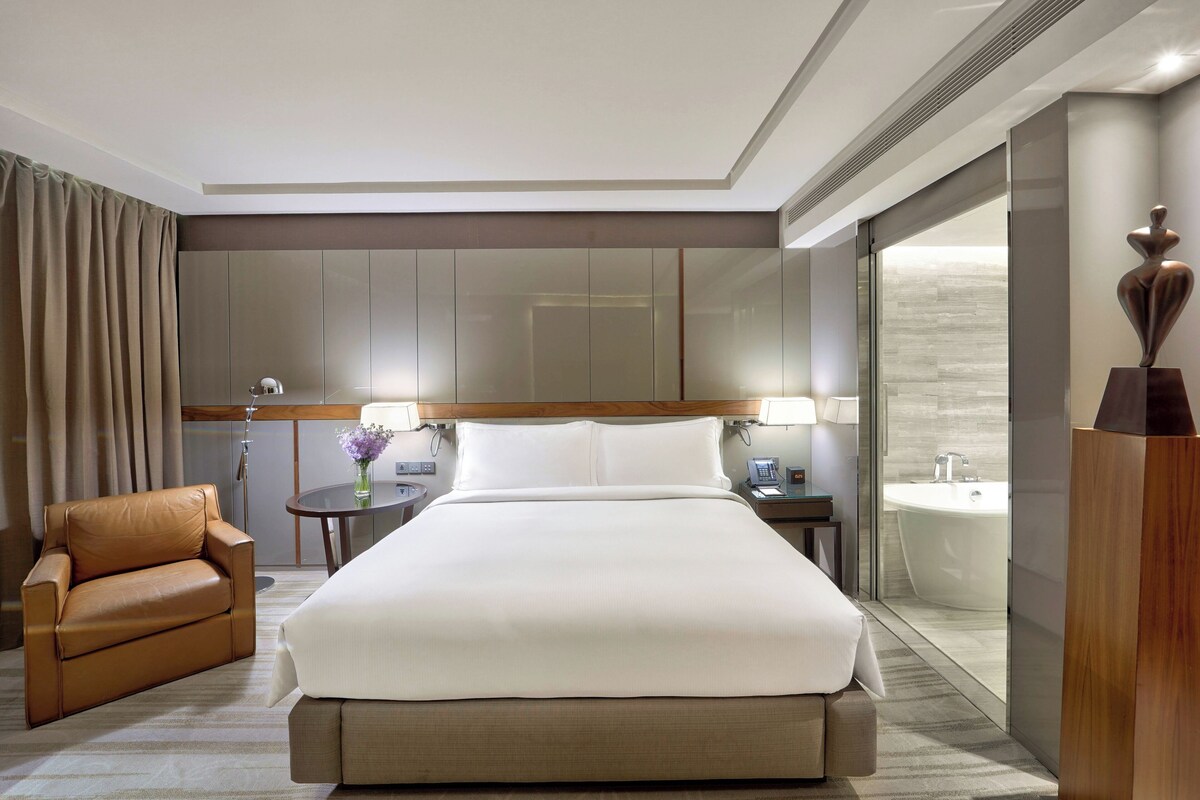 Une chambre d’hôtel avec un grand lit, un fauteuil en cuir, une table de chevet avec une lampe, un miroir et une vue sur une salle de bain avec une baignoire.