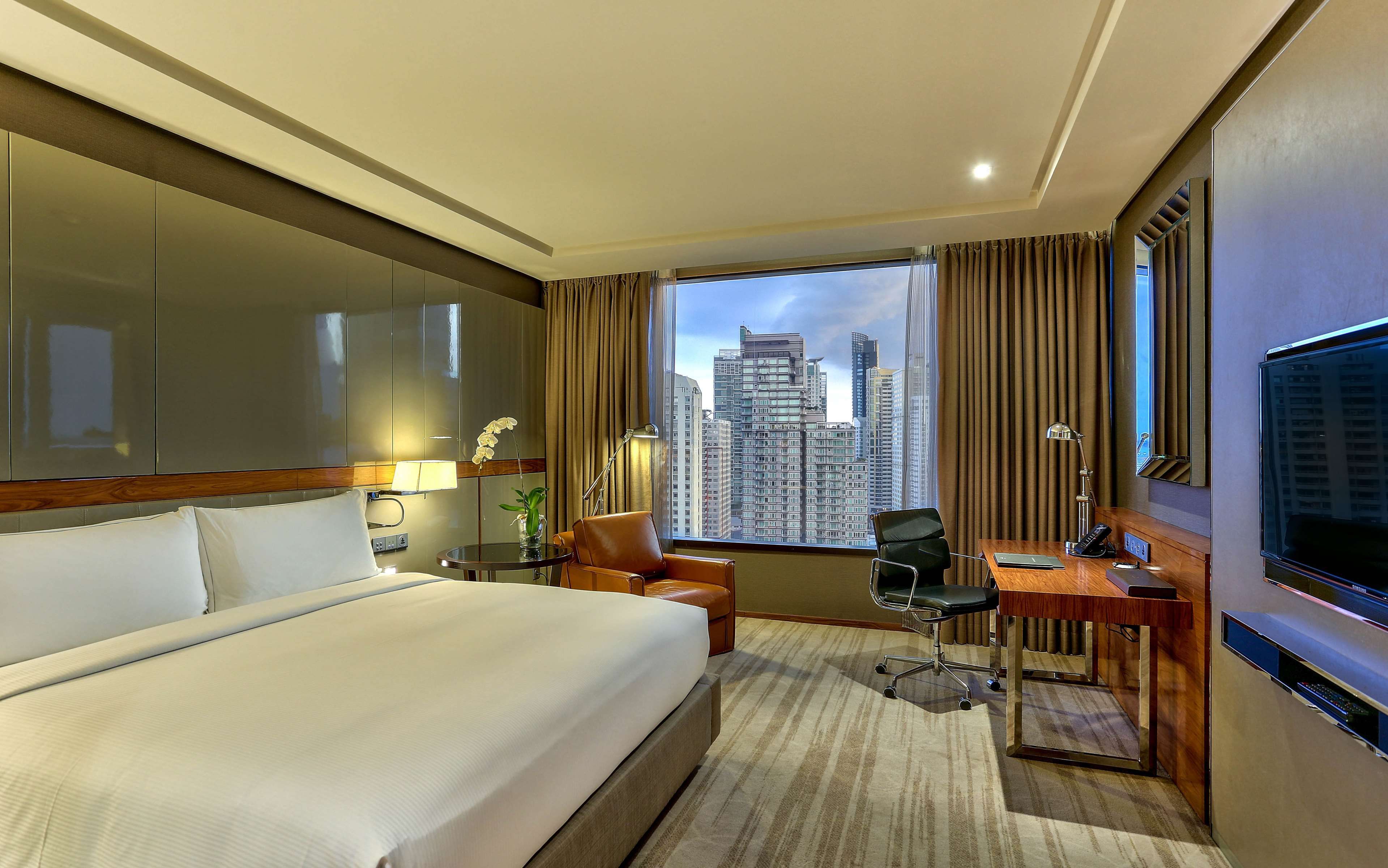 A hotel room with a large bed, a desk with a chair, a TV, and a view of tall buildings.