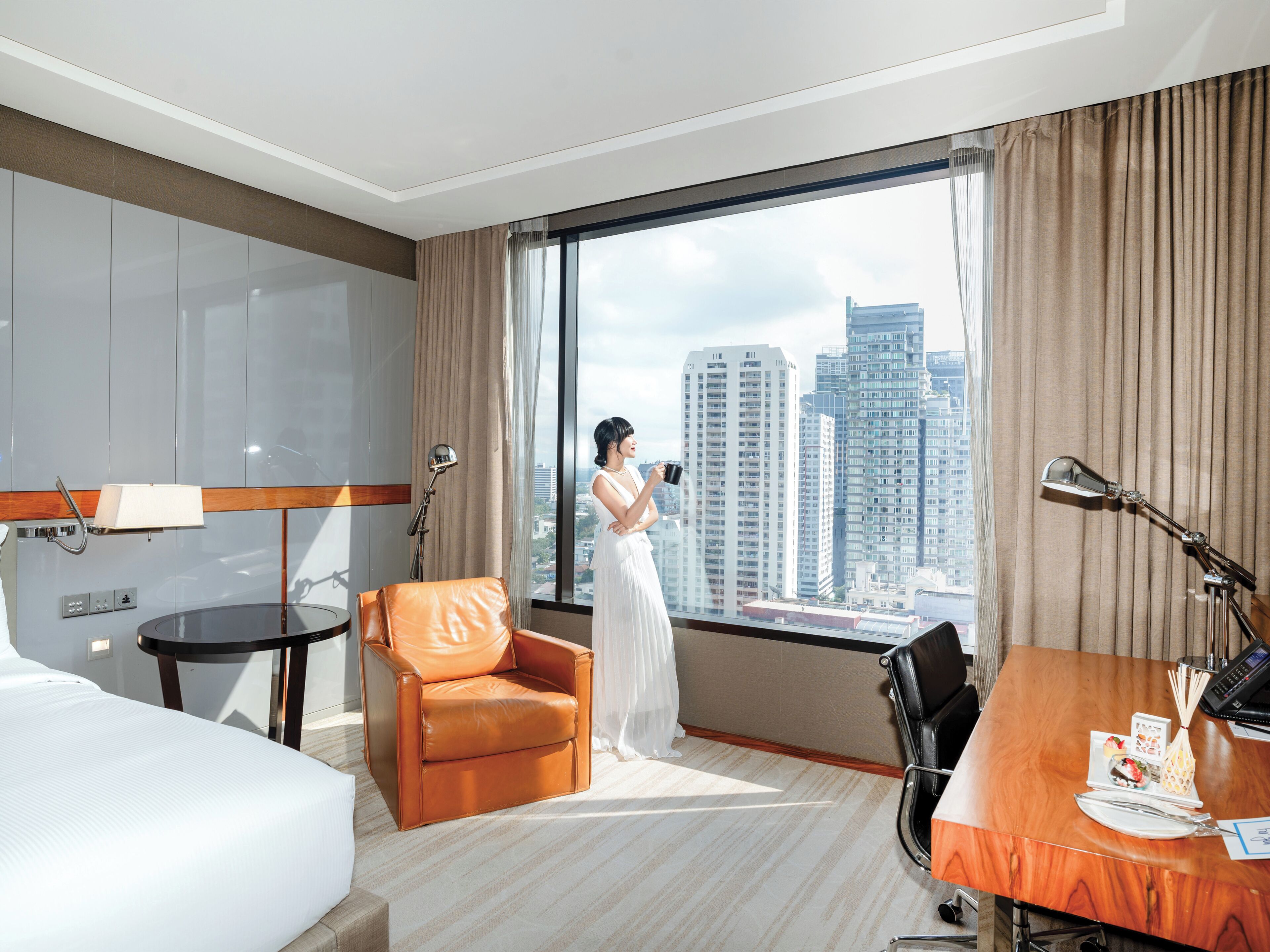 A hotel room with a large window, a bed, a desk, a chair, and a lamp.