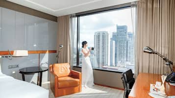 A hotel room with a large window, a bed, a desk, a chair, and a lamp.