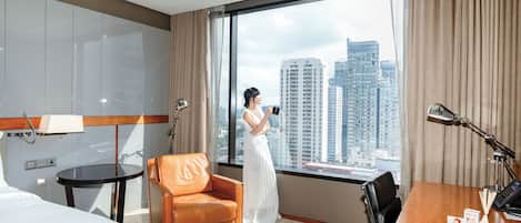 A hotel room with a large window, a bed, a desk, a chair, and a lamp.
