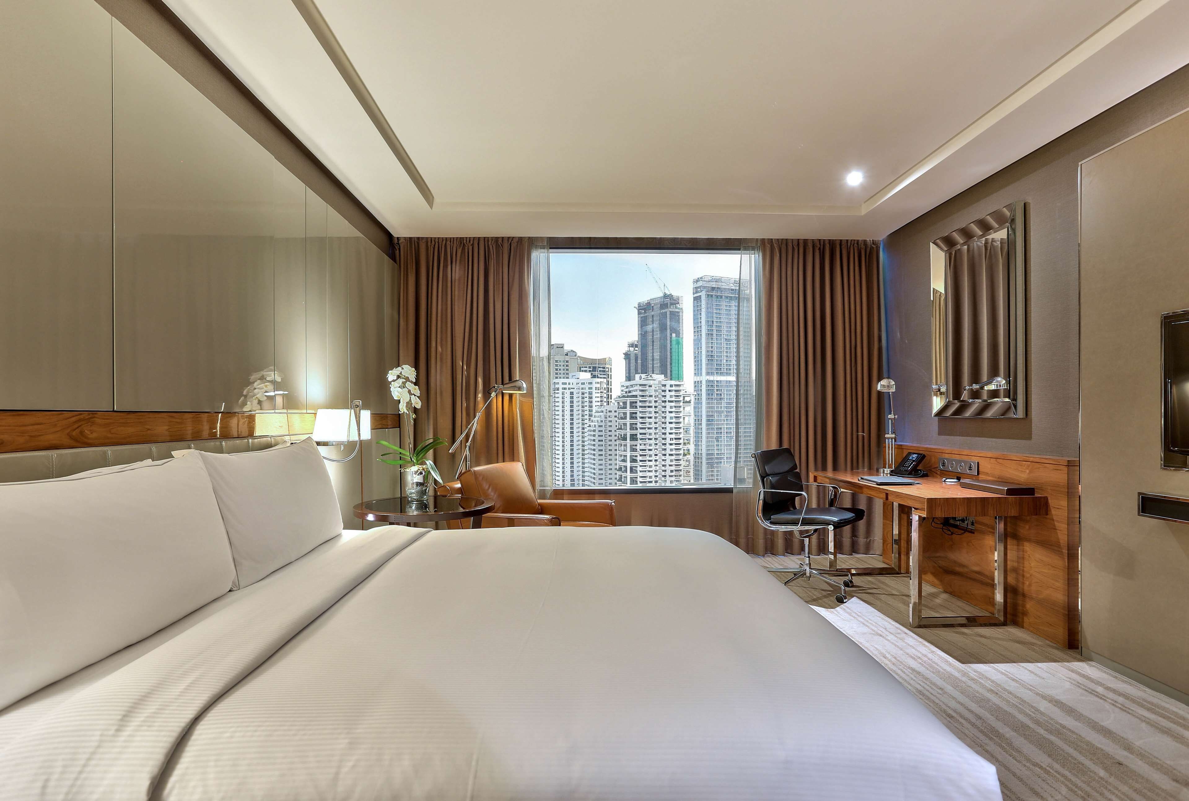 A hotel room with a large bed, a desk, a chair, a mirror, and a view of tall buildings.
